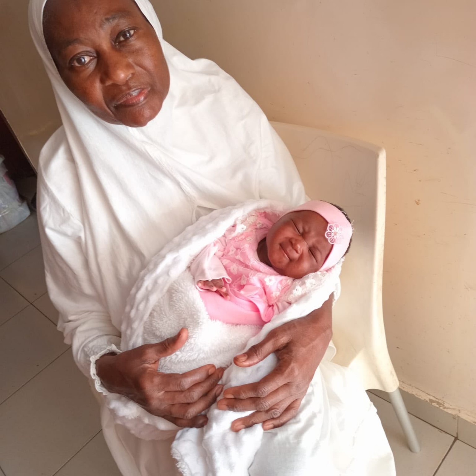 A grandmother in a white headscarf holding her newborn granddaughter, who is wrapped in a pink outfit and a white blanket.